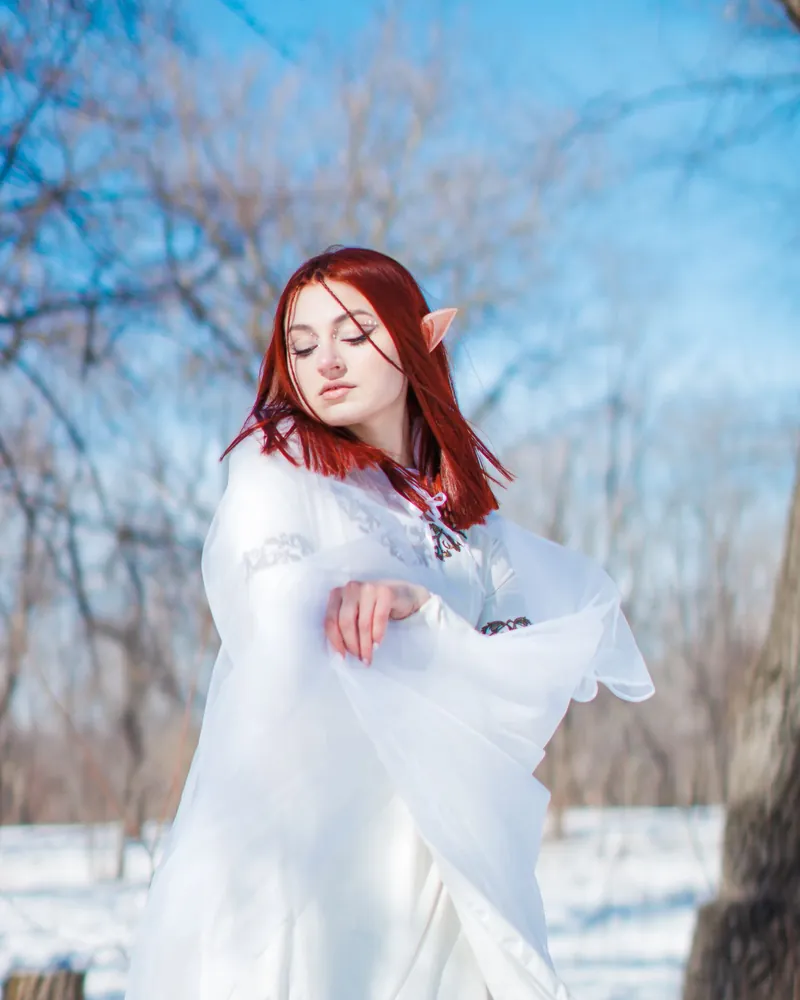 Woman in an elf costume with red hair and a white dress in a snowy forest